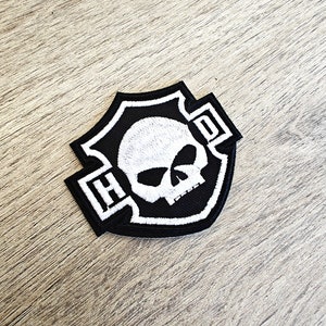 Könnte beinhalten: Schwarz-weiß gestickter Patch mit Totenkopf- und Knochenkreuz-Motiv. Der Patch zeigt die Buchstaben "HD" in einer stilisierten Schrift.