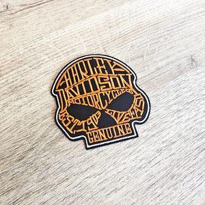 Può includere: Toppa a forma di teschio Harley-Davidson nera e arancione. La toppa presenta il logo Harley-Davidson e le parole "EST. 1903 GENUINE" in un carattere arancione stilizzato. La toppa ha un bordo bianco.