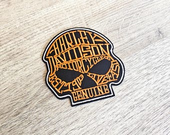May include: Black and orange Harley-Davidson skull patch. The patch features the Harley-Davidson logo and the words "EST. 1903 GENUINE" in an orange, stylized font. The patch has a white border.