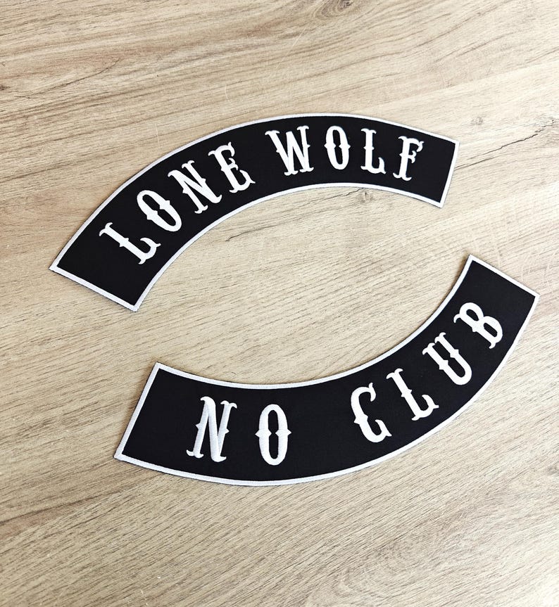 May include: Two black and white arched patches. The top patch reads "LONE WOLF" in white, stylized lettering. The bottom patch reads "NO CLUB" in white, stylized lettering. The patches are on a light brown wooden surface.