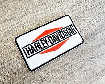 May include: A rectangular white patch featuring the text "HARLEY-DAVIDSON" in black, framed by a black border and two red arrow motifs. The patch is designed for application on clothing.