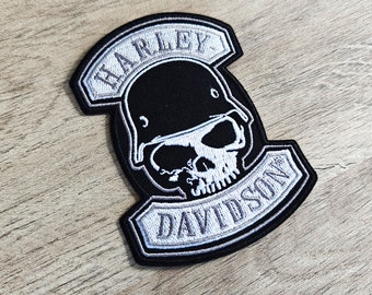 May include: A black and white Harley-Davidson patch featuring a skull wearing a helmet. The patch has the words "HARLEY" and "DAVIDSON" in a stylized font. The patch is on a wooden surface.