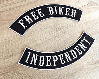 May include: Two black, arched patches with white text. The top patch reads "FREE BIKER" and the bottom patch reads "INDEPENDENT". Both patches have a white border. The patches are on a light brown wooden surface.