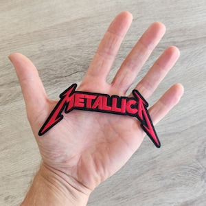 Op de afbeelding: Een rood en zwart geborduurd embleem met het woord "METALLICA" in een gestileerd lettertype. Het embleem heeft een zwarte rand en de vorm van het logo van de band. Het is bedoeld om op kleding of accessoires te naaien of te strijken.