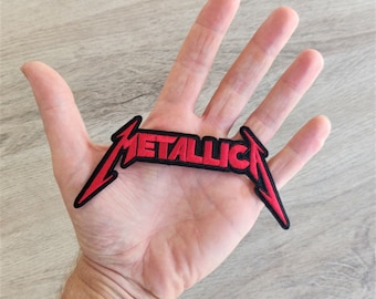 May include: A red and black embroidered patch with the word "METALLICA" in a distinctive font. The patch has a black border and is in the shape of the band's logo. It is designed to be sewn or ironed onto clothing or accessories.