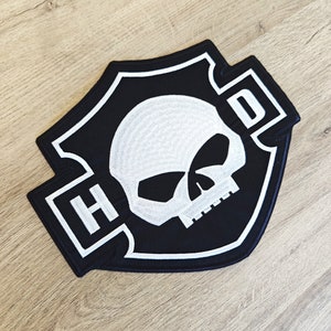 Puede incluir: Parche bordado en blanco y negro con un diseño de calavera y huesos cruzados. El parche presenta las letras "HD" en un contorno blanco.