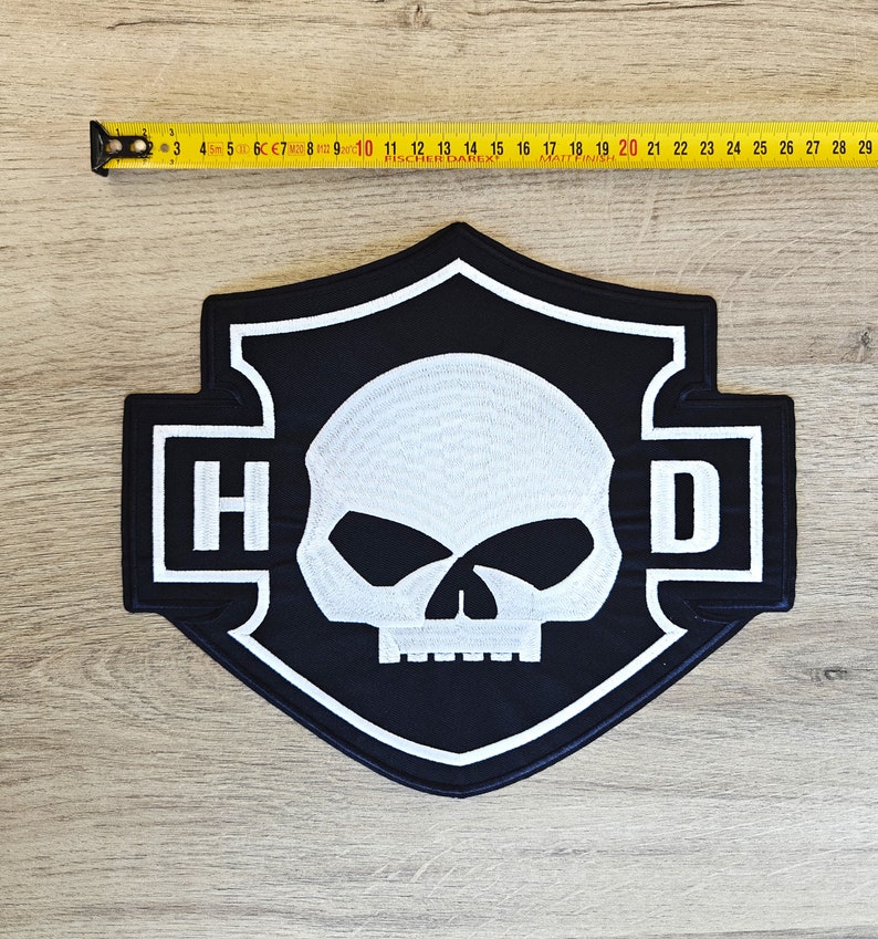 May include: Black and white embroidered patch with a skull and crossbones design. The patch features the letters "H-D" on either side of the skull.