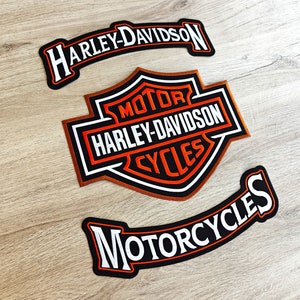 Puede incluir: Tres parches bordados negros y naranjas con letras blancas. El parche más grande presenta un logotipo de Harley-Davidson con las palabras "Motor", "Harley-Davidson" y "Cycles". Los otros dos parches son curvos y dicen "Harley-Davidson" y "Motorcycles".