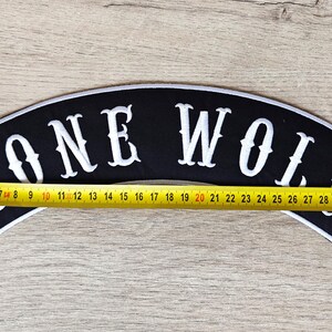 May include: Black, arched patch with the words "LONE WOLF" in white, outlined in white. The patch is approximately 13.75 inches long. A yellow measuring tape is placed across the patch.