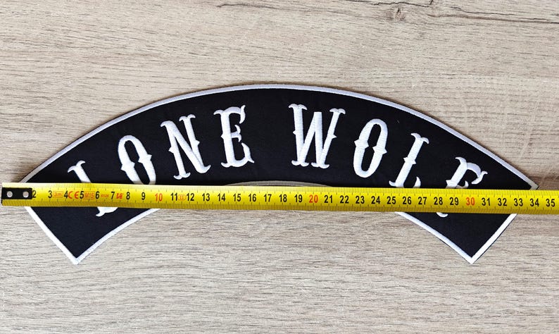 May include: Black, arched patch with the words "LONE WOLF" in white, outlined in white. The patch is approximately 13.75 inches long. A yellow measuring tape is placed across the patch.