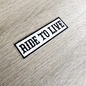 Könnte beinhalten: Ein rechteckiger, weißer Aufnäher mit den Worten "RIDE TO LIVE" in Schwarz gestickt, umrandet von einem schwarzen Rand. Der Aufnäher wird auf einer hellen Holzmaserungsoberfläche gezeigt.