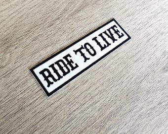 May include: A rectangular white patch with the words "RIDE TO LIVE" embroidered in black, outlined with a black border. The patch is displayed on a light wood-grain surface.