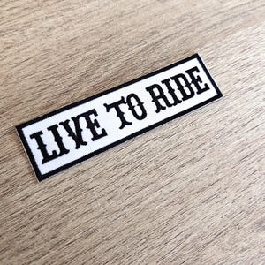 Könnte beinhalten: Ein rechteckiger Aufnäher mit dem Text "LIVE TO RIDE" in fetter, schwarzer, stilisierter Schrift. Der Text befindet sich auf weißem Grund mit schwarzem Rand. Der Aufnäher ist für Kleidung oder Accessoires bestimmt.