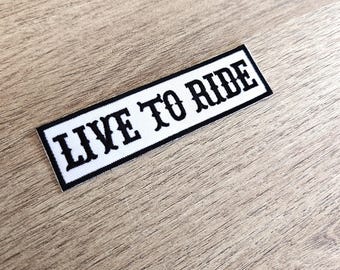 May include: A rectangular patch featuring the text "LIVE TO RIDE" in bold, black lettering. The words are on a white background with a black border. The patch is designed for application on clothing or accessories.