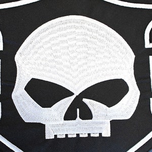 May include: Black and white embroidered patch with a skull design. The skull is white with black eyes and a black mouth. The patch is shaped like a shield with a white border.