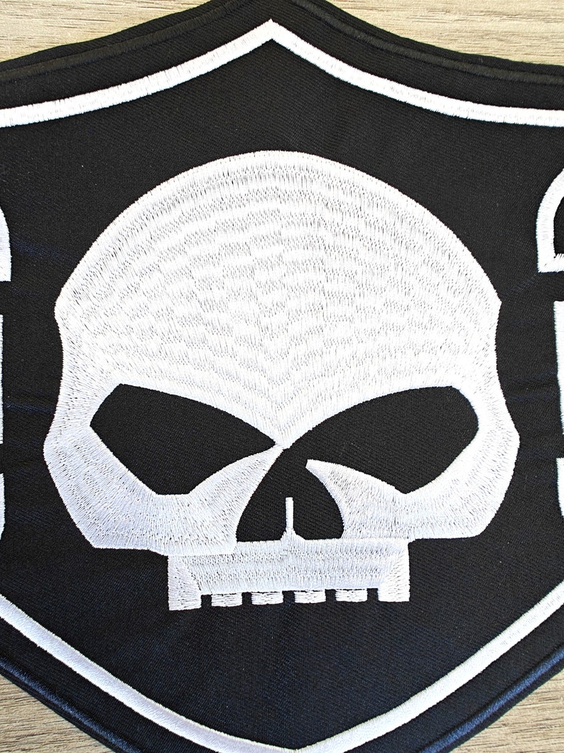 May include: Black and white embroidered patch with a skull design. The skull is white with black eyes and a black mouth. The patch is shaped like a shield with a white border.