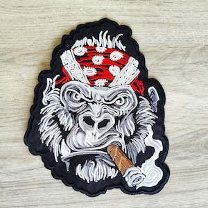 Puede incluir: Parche bordado para planchar con un gorila gris que lleva un pañuelo rojo con lunares blancos y fuma un cigarro marrón. El gorila tiene una expresión seria y está rodeado de un fondo negro.