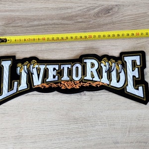 Può includere: Toppa ricamata nera con scritta bianca che recita "Live to Ride". Le lettere sono contornate d'oro e hanno un segno di artiglio dorato su entrambi i lati. C'è un disegno di filo spinato dorato con una rosa rossa al centro.