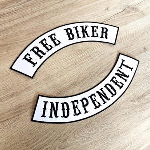 Könnte beinhalten: Zwei weiße, gebogene Aufnäher mit schwarzem Text. Der obere Aufnäher lautet "FREE BIKER" und der untere Aufnäher "INDEPENDENT". Die Aufnäher haben einen schwarzen Rand und liegen auf einer hellbraunen Holzoberfläche.