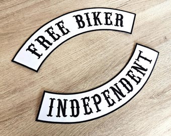 May include: Two white, curved patches with black text. The top patch says "FREE BIKER" and the bottom patch says "INDEPENDENT". The patches have a black border and are on a light brown wooden surface.