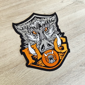 Può includere: Toppa ricamata raffigurante una testa di cinghiale con occhi e zanne arancioni. La toppa presenta la scritta "HOG" in arancione e giallo, con fiamme. La toppa è bordata di nero.
