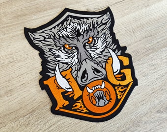May include: Embroidered patch depicting a wild boar's head with orange eyes and tusks. The patch features the word "HOG" in orange and yellow, with flames. The patch is outlined in black.