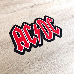 Peut inclure: Un écusson noir avec le texte rouge "AC/DC" dans une police stylisée, avec un éclair au centre. Le texte est bordé de blanc. L'écusson est posé sur une surface en bois clair.