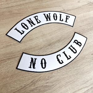 Op de afbeelding: Twee witte, gebogen patches met zwarte tekst. De bovenste patch leest "LONE WOLF" en de onderste patch leest "NO CLUB". De patches hebben een zwarte rand en worden weergegeven op een lichtbruin houten oppervlak.