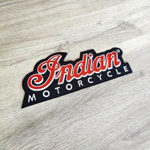 Może przedstawiać: Czarna naszywka z napisem "Indian MOTORCYCLE" w białych i czerwonych literach. Słowo "Indian" jest w stylizowanej czcionce. Naszywka ma czarną obwódkę i znajduje się na jasnej powierzchni z usłojeniem drewna.