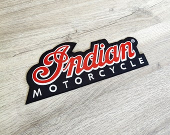 May include: A black patch featuring the words "Indian MOTORCYCLE" in white and red. The word "Indian" is in a decorative script. The patch has a black border and is set against a light wood grain background.