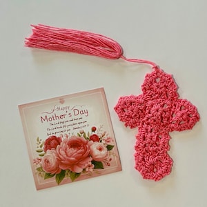 May include: A pink crocheted cross with a tassel, next to a Mother's Day card. The card features a floral design with the text "Happy Mother's Day" and a Bible verse. The cross is made of pink yarn.
