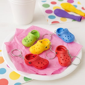 May include: A collection of colorful miniature clogs keychains in green, yellow, red, and blue, arranged on a pink paper napkin. The keychains are on a white plate, with colorful polka dot napkins and utensils in the background.