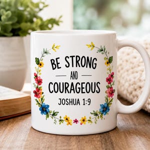 May include: A white ceramic mug featuring a floral wreath design in shades of red, blue, and yellow. The text in the center reads, "BE STRONG AND COURAGEOUS JOSHUA 1:9" in a black sans-serif font. The mug has a curved handle on the right side.