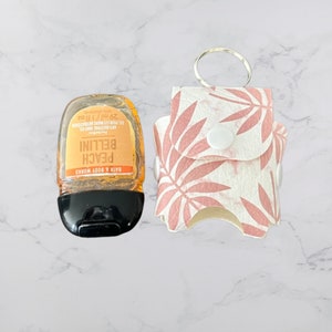 以下が含まれることがあります： ピンクと白の花柄のハンドサニタイザーホルダー。スナップ留めとシルバーのキーリングが付いています。ホルダーは、Bath & Body Worksのピーチ・ベリニハンドサニタイザーの小さなボトルに取り付けられています。