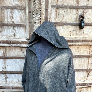 May include: A gray hooded coat with a herringbone pattern. The coat has a large hood and a relaxed fit. The coat is displayed against a weathered wooden door with metal accents and a black padlock.