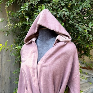 May include: A mauve hooded robe with a matching belt tied at the waist. The robe has long sleeves and a soft, textured appearance. The hood is lined with a lighter shade of mauve.
