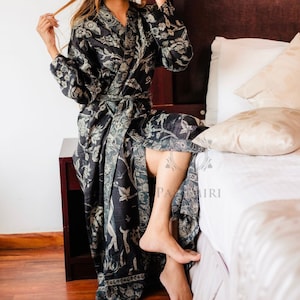 May include: A dark grey robe featuring a floral and animal pattern, secured with a matching belt. The robe has long sleeves and falls to the knees. The person is seated on a dark brown wooden surface next to a bed with white bedding and pillows.