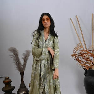 May include: An olive green robe with a white and grey floral pattern, worn by a person. The robe has wide sleeves and a matching belt. The background features vases, rugs, and dried plants.