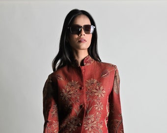 May include: A red and gold floral patterned jacket with a mandarin collar and button closure. The jacket is paired with tan trousers. The person is wearing sunglasses.