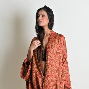 May include: A full-length shot of a red and gold patterned kimono-style robe. The robe has a floral design and is draped over the body. The sleeves are wide and the robe is open in the front.