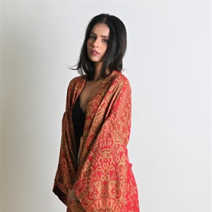 May include: A long, flowing red and gold patterned kimono-style robe. The robe features an intricate paisley design and wide sleeves. The model is wearing a black top underneath. The robe is floor-length.