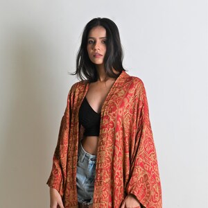 May include: A long, open-front robe in shades of red and gold, with an intricate paisley pattern. The robe has long sleeves and pockets. The fabric appears lightweight and flowing. The garment is worn over a black top and denim shorts. The robe is suitable for layering.