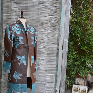 May include: A brown duster coat with teal bird and floral patterns displayed on a wooden mannequin. The coat has long sleeves and a collarless design. The mannequin is on a dark wooden stand, set against a backdrop of a bamboo blind and a weathered wooden door.