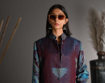 May include: A long, button-down jacket in burgundy and teal, with a crane design. The jacket has a mandarin collar and pockets. A dark blue handbag with gold hardware is held. The person is wearing sunglasses.