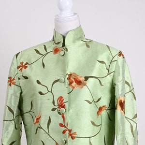 May include: A light green jacket with a mandarin collar and floral embroidery. The jacket features a button-up front and two small pockets. The floral design includes orange and brown flowers with green stems and leaves.