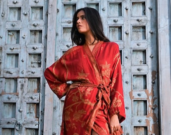 May include: A long, red robe with a floral pattern in gold. The robe is tied at the waist and has long sleeves. The model is wearing clear heels. The background is a weathered, blue wooden door.