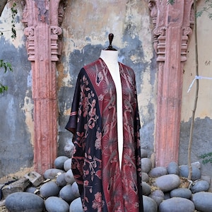 May include: A long, open-front kimono-style robe in black and burgundy. The robe features a floral and bird pattern. The garment is displayed on a mannequin in an outdoor setting with stone and architectural elements in the background.