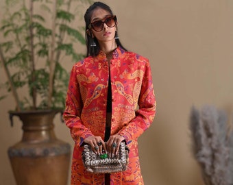 May include: A long, open-front coat in shades of red, orange, and purple, featuring a peacock pattern. The coat is worn with dark denim jeans and a small, embellished handbag. The coat's design includes a mandarin collar.