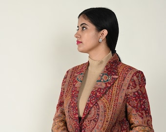 May include: A burgundy blazer with an intricate paisley pattern in shades of gold, blue, and green. The blazer is worn over a tan turtleneck and blue jeans. The jacket has a classic tailored fit.