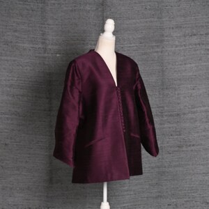 May include: A plum-colored jacket with a V-neck and three buttons, displayed on a white mannequin. The jacket has long sleeves and two front pockets. The mannequin is on a white stand with a three-legged base.
